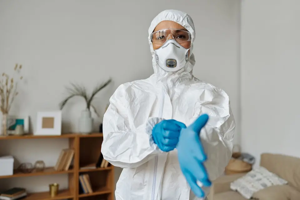 Personne en combinaison blanche à capuche, lunettes et masque respiratoire, enfilant ses gants dans une pièce d'habitation